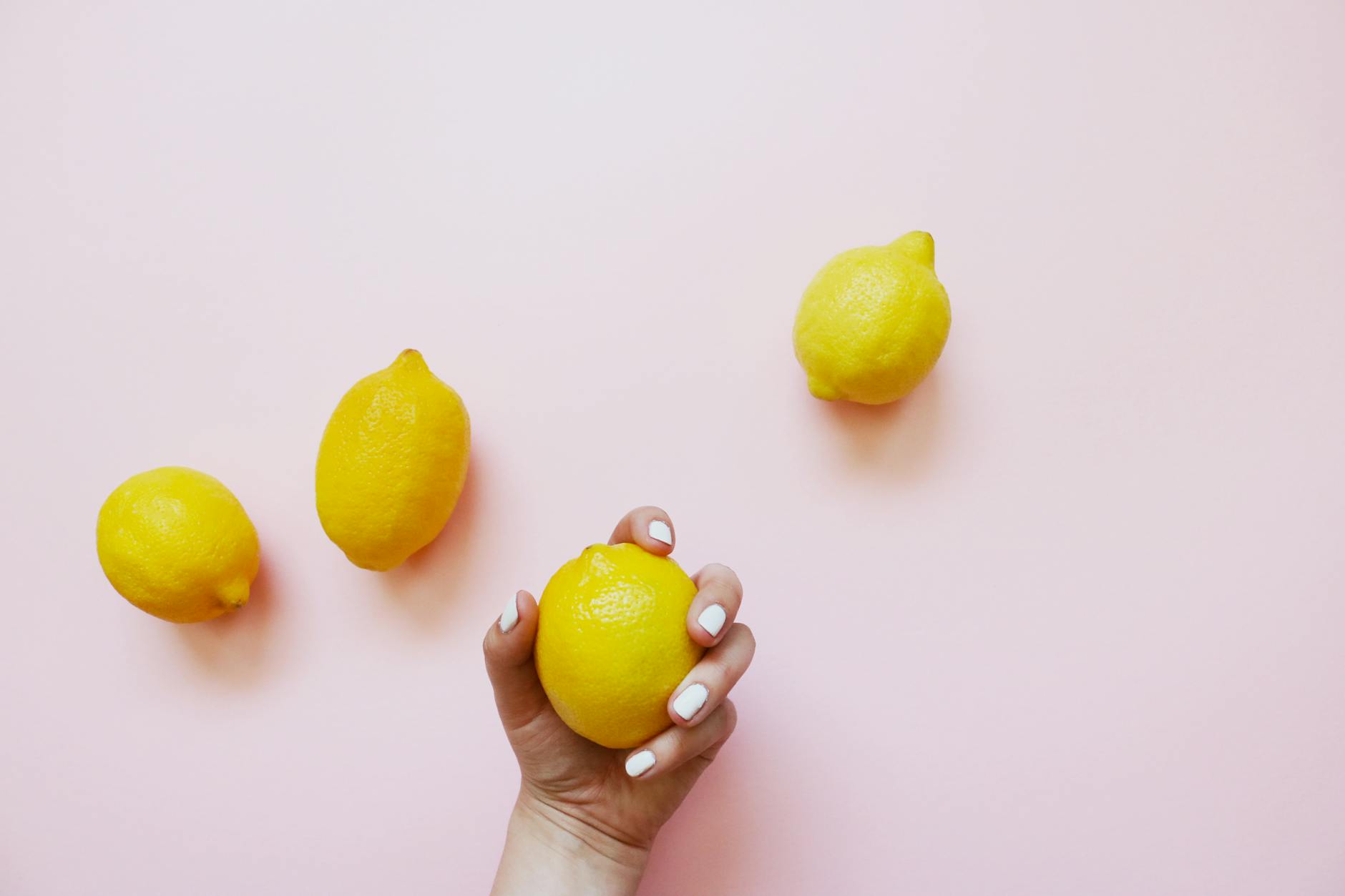 A hand with white nails gently holding a lemon on a soft pink background, symbolizing self-care and gentle healing.