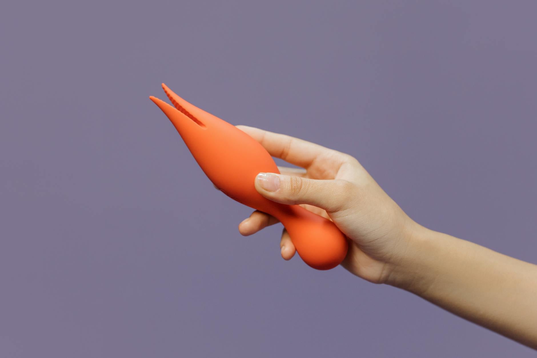 Eine Hand hält einen orangefarbenen Vibrator gegen einen minimalistischen lila Hintergrund